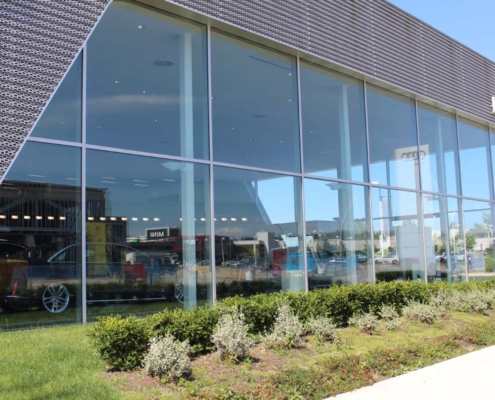 Large glass windows of a modern car dealership reflect the sky and nearby buildings, with several cars visible inside. There are green bushes and grass landscaping in the foreground.