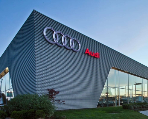 A modern Audi dealership building with large glass windows and metallic exterior, displaying the Audi logo and name on the upper facade. Bushes and grass are in the foreground under a clear sky.