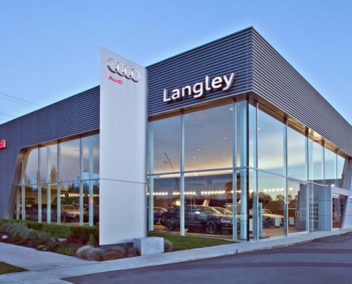 Modern Audi dealership building with large glass windows, metallic exterior, and illuminated Audi and Langley signs at dusk. Several cars are visible inside the brightly lit showroom.