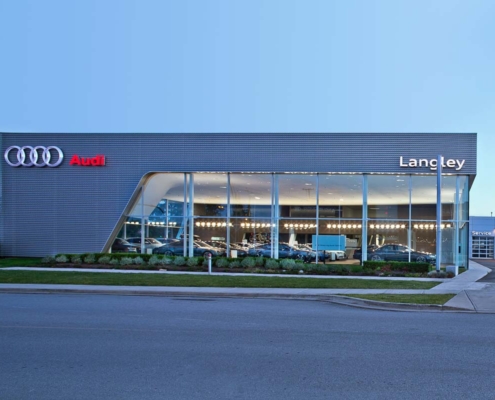 A modern Audi dealership building with large glass windows, displaying several cars inside. The signs read “Audi” in red and “Langley” in white. The exterior is sleek, with a manicured landscape and a clear sky overhead.