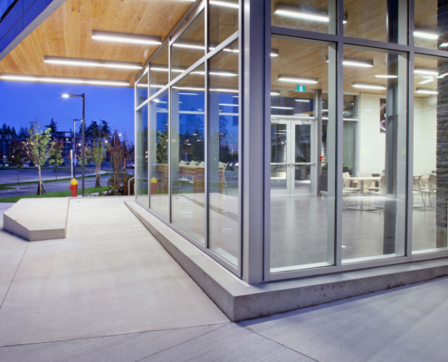 Modern building with large glass windows and a wooden ceiling, showing an empty, well-lit interior with tables and chairs at dusk. The exterior has a concrete sidewalk and landscaping with trees.