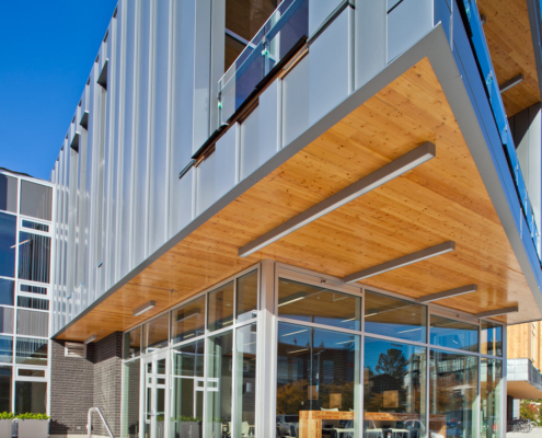 A modern building with large glass windows and metal siding features an overhanging section with a wooden ceiling. The entrance below has glass doors and visible interior seating. Blue sky and trees are in the background.