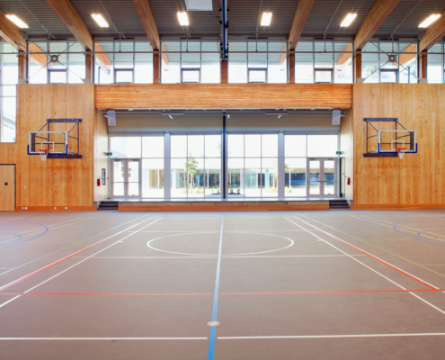 A spacious indoor gymnasium with a wooden interior, two basketball hoops on opposite walls, large windows letting in natural light, and marked courts on the floor.