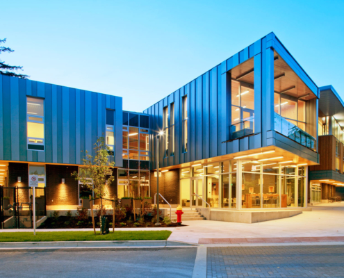 Modern building with large glass windows and blue metal panels, illuminated at dusk. Trees and landscaping surround the structure, and warm interior lights are visible through the windows. A red fire hydrant is at the curb.