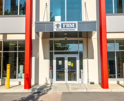 Entrance to a modern building with red columns and a glass door. A sign above the door reads FBM Foundation Building Materials. Yellow bollards are positioned near the entrance.