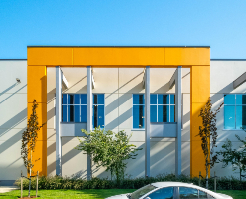 A modern building with a white and yellow facade, tall vertical columns, large windows, and a white car parked in front. Trees and green grass are visible in the foreground under a clear blue sky.