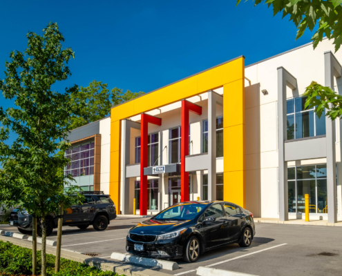 A modern office building with a bold yellow and red entrance facade, large windows, and parked cars in the lot on a sunny day. Trees and greenery are visible around the parking area.