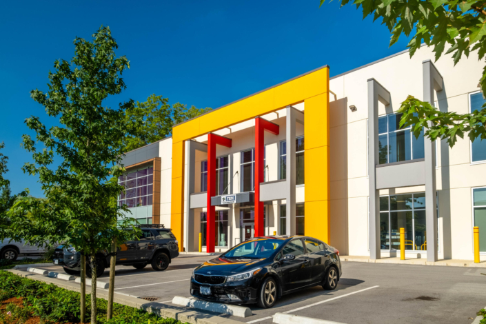 A modern office building with a bold yellow and red entrance facade, large windows, and parked cars in the lot on a sunny day. Trees and greenery are visible around the parking area.